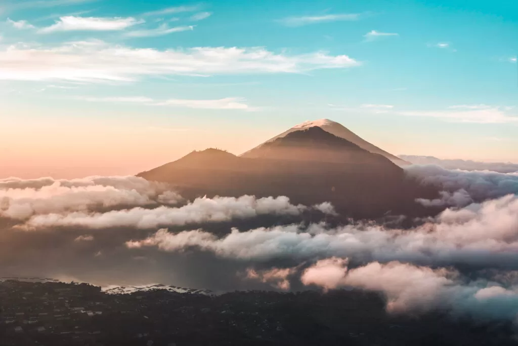 This photo shows the top of mount Batur at sunrise. The sky is blue and the sun spreads a pink hue. There are clouds surrounding the volcano, giving it a dreamy look.