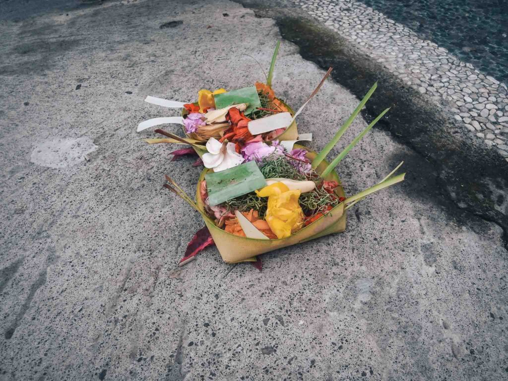 This photo shows a traditional Balinese offering basket. It is made from leaves and inside is rice, fruit, herbs, flowers and incense. It is lying on a paved sidewalk.