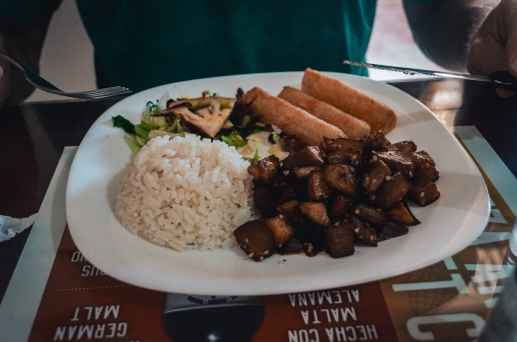 A plate of traditional Colombian lunch featuring a scoop of white rice, golden-brown diced eggplants, crispy yukka fries, and a side salad, set on a table with a diner holding utensils, ready to eat.