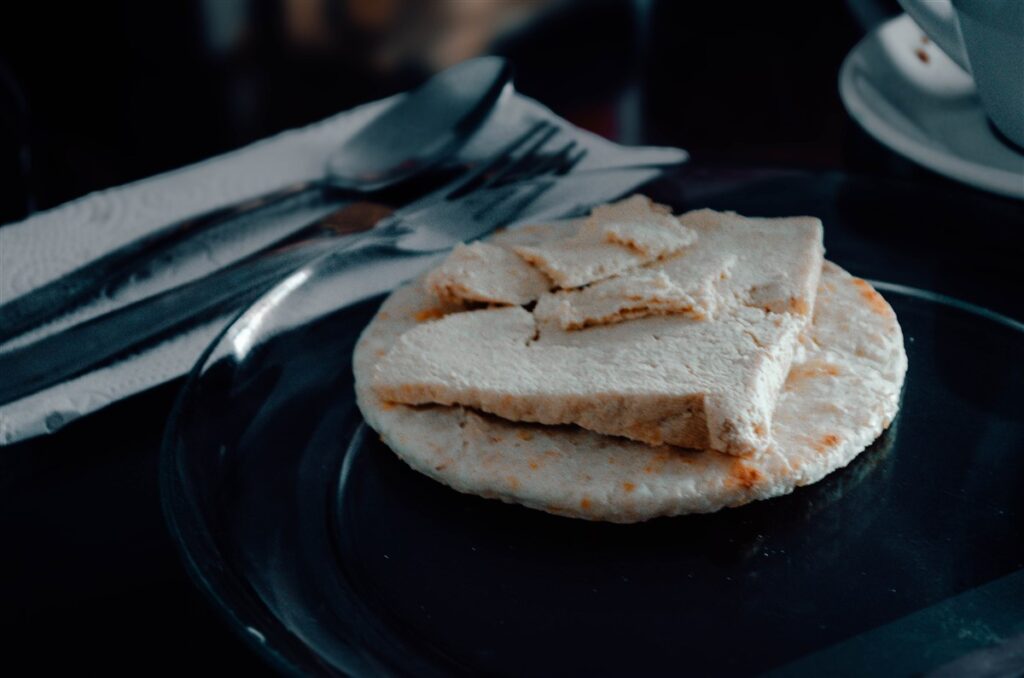 This photograph showcases a vegan twist on a quintessential Colombian dish. We see a well-toasted arepa, a staple of Colombian cuisine, topped with a generous slice of tofu cheese, suggesting a mindful substitution for those following a plant-based diet. The arepa's surface is lightly speckled with golden spots, indicative of being freshly grilled or pan-fried. The tofu cheese, with its creamy hue and firm texture, sits in stark contrast to the dark ceramic plate beneath it. This simple yet hearty combination is an excellent representation of how traditional Colombian food can be adapted to suit vegan preferences. The table setting is complete with a folded white paper napkin and silverware on the side, all set against a subdued background, focusing all attention on the meal.