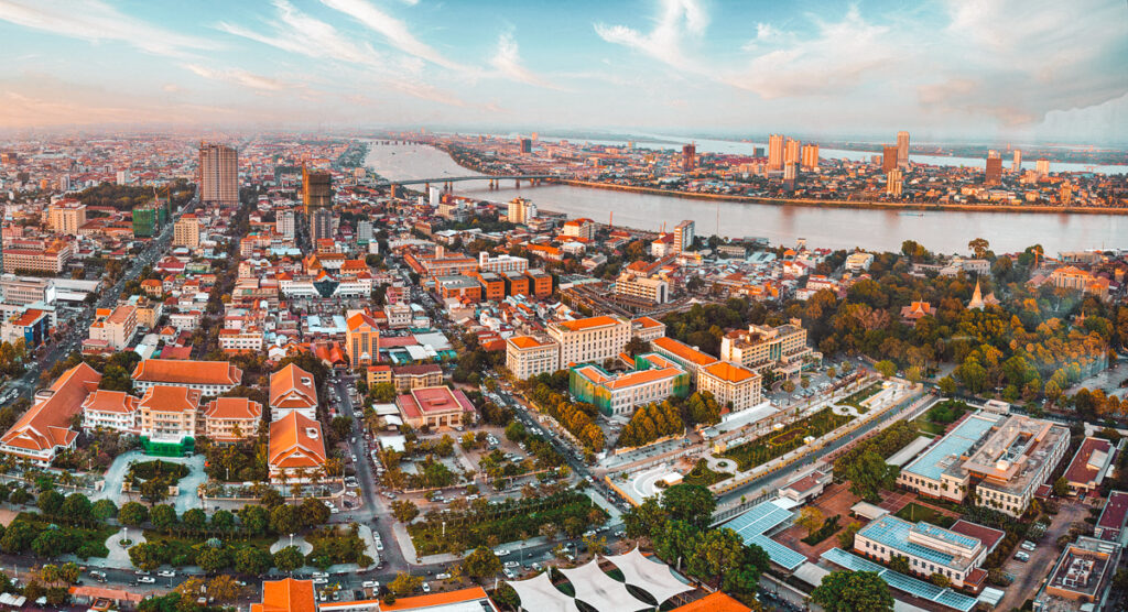 Aerial view of Phnom Penh, the capital city of Cambodia, showcasing a mix of traditional terracotta-roofed buildings and modern skyscrapers, with a prominent river winding through the urban landscape during the golden hour.