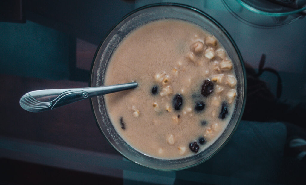 A clear bowl of vegan Colombian sweet mute soup containing corn kernels and raisins. The beige-colored broth consist of nut milk and cinnamon. A single metal spoon is placed in the soup, which sits on a reflective glass surface. The environment is faintly mirrored on the table, adding to the photo's depth. This image represents how Colombian cuisine can be adapted for a vegan diet.