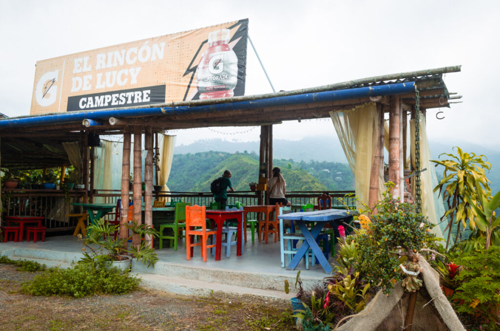 A rustic outdoor seating area at 'El Rincón de Lucy Campestre', a casual dining spot in Salento, Colombia. The place features a mix of colorful wooden chairs and tables under a simple roof with bamboo pillars. A large advertisement banner for a beverage is displayed prominently above. Two people are seen standing at the railing, enjoying the misty mountain view in the background, amidst lush greenery. The scene is adorned with vibrant potted plants and a draped fabric, giving it a cozy and inviting atmosphere