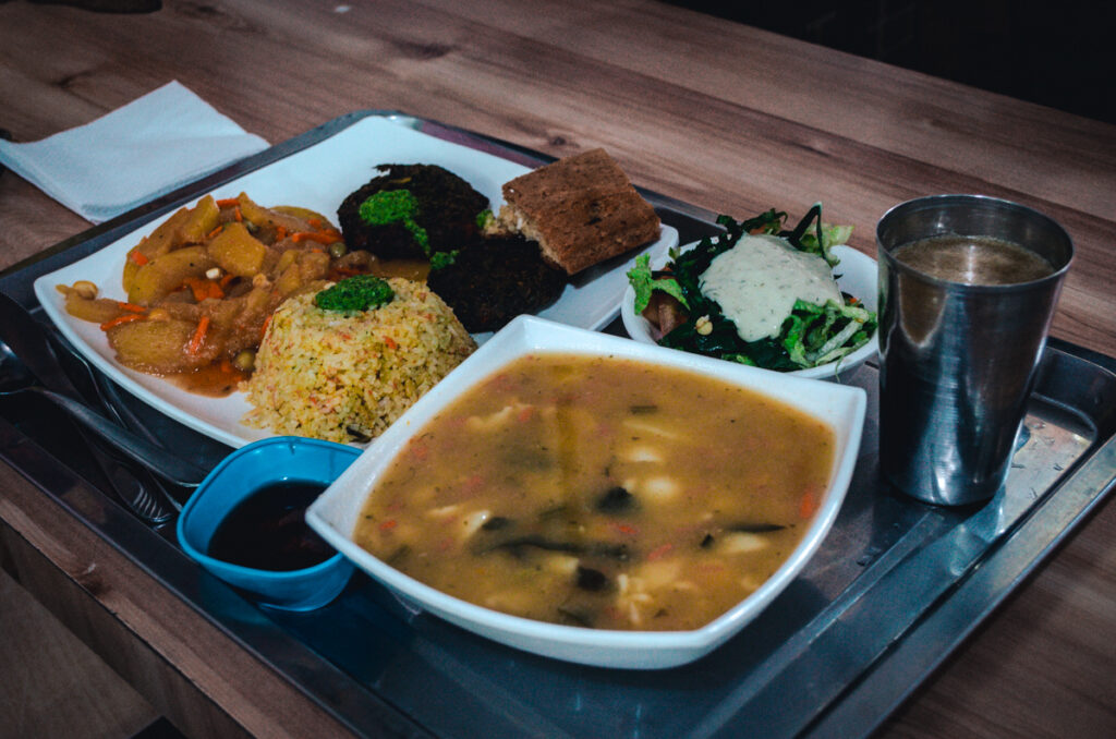 A vibrant, well-balanced tray of vegan Colombian food from Govindas restaurant in Bogotá, Colombia. The meal includes a variety of colorful dishes: a side of stewed vegetables, a serving of yellow seasoned rice, dark veggie patties, a fresh green salad topped with a creamy dressing, and a hearty bowl of vegetable soup. Accompanying the meal is a piece of cake and a metal cup with juice. The meal is neatly arranged on a metal tray, ready to be enjoyed in a casual dining setting.
