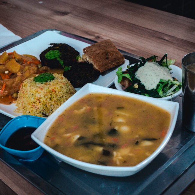 A vibrant, well-balanced tray of vegan Colombian food from Govindas restaurant in Bogotá, Colombia. The meal includes a variety of colorful dishes: a side of stewed vegetables, a serving of yellow seasoned rice, dark veggie patties, a fresh green salad topped with a creamy dressing, and a hearty bowl of vegetable soup. Accompanying the meal is a piece of cake and a metal cup with juice. The meal is neatly arranged on a metal tray, ready to be enjoyed in a casual dining setting.