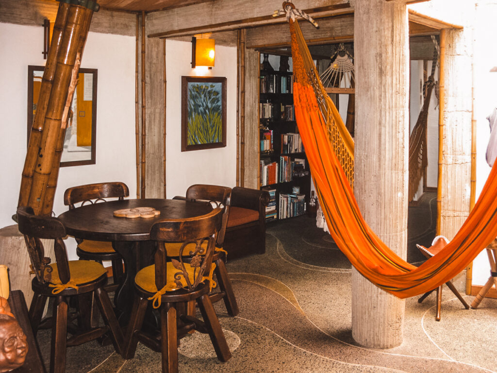 A cozy dining area in the main house at Kasaguadua Natural Reserve, featuring a round wooden table with matching chairs and cushions. A vibrant orange hammock is strung between two sturdy columns, adding a relaxing touch to the room. Bamboo elements, bookshelves, and warm lighting create a welcoming and homely atmosphere.