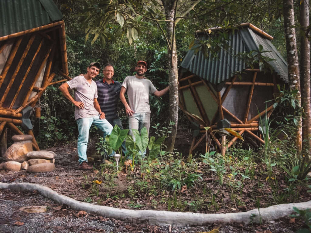 members of the Kasaguadua crew stand proudly on a path in the Kasaguadua Natural Reserve, Salento. Behind them are two distinctive hexagonal sleeping pods built on stilts, harmoniously integrated into the dense and verdant forest environment, showcasing the eco-friendly accommodations offered at the reserve.