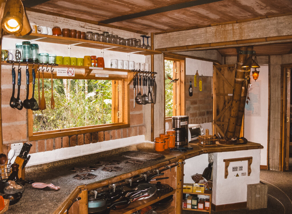 The rustic kitchen in the main house of Kasaguadua Natural Reserve is equipped with traditional cookware and a variety of colorful mugs hanging on the shelves. A large window opens to the surrounding greenery, allowing natural light to fill the space. Wooden countertops and tools echo the naturalistic ethos of the reserve.