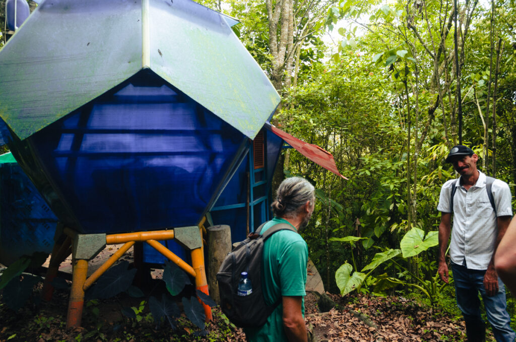 Visitors on a tour at Kasaguadua Natural Reserve are engaging with a local guide near an eco-friendly, hexagonal blue sleeping pod raised on stilts. The structure is set amidst lush greenery, with large leaves and trees surrounding the area, suggesting a deep immersion in the natural environment of the reserve.