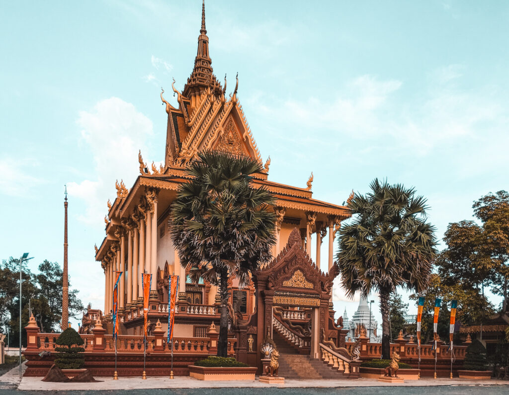 A traditional Khmer temple in Phnom Penh, Cambodia, with ornate golden spires and intricate carvings, flanked by lush palm trees under a clear blue sky.