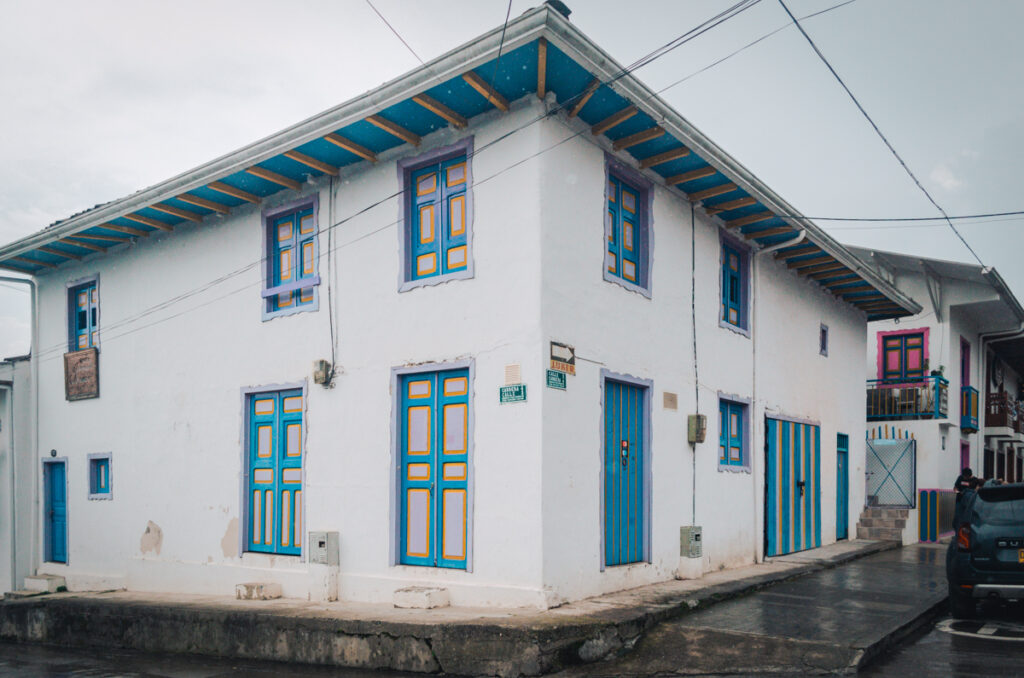 A two-story corner building in Salento, Colombia, with white walls and bright blue trimmed windows and doors. A sign on the upper floor reads 'Jerico Hostal Familiar.' The sky is overcast, and the streets are wet, suggesting recent rain