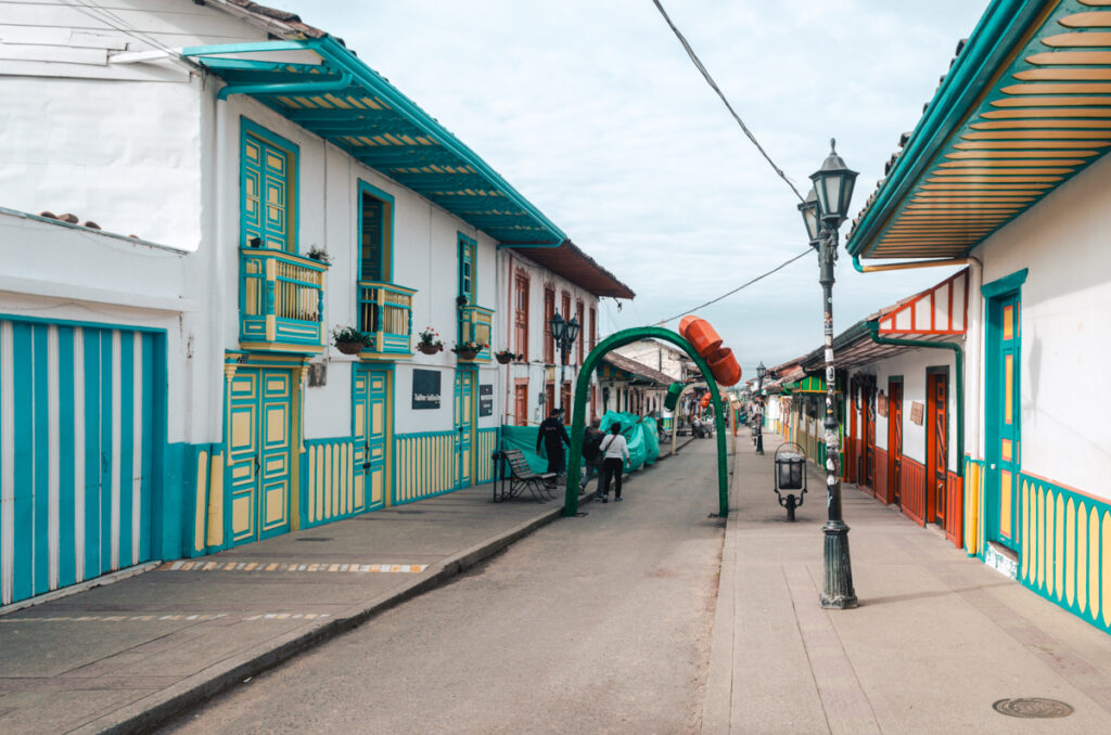 A vibrant street scene in Salento, Colombia, showcasing the town's charming colonial architecture. Colorful buildings with blue and green trim line the cobblestone street, with ornate balconies adorned with flowers. Traditional lampposts stand along the sidewalk, and a series of green arches run down the street as christmas decorations. The sky is overcast, and a few people can be seen enjoying the tranquil atmosphere of this quaint town.