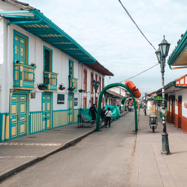 A vibrant street scene in Salento, Colombia, showcasing the town's charming colonial architecture. Colorful buildings with blue and green trim line the cobblestone street, with ornate balconies adorned with flowers. Traditional lampposts stand along the sidewalk, and a series of green arches run down the street as christmas decorations. The sky is overcast, and a few people can be seen enjoying the tranquil atmosphere of this quaint town.