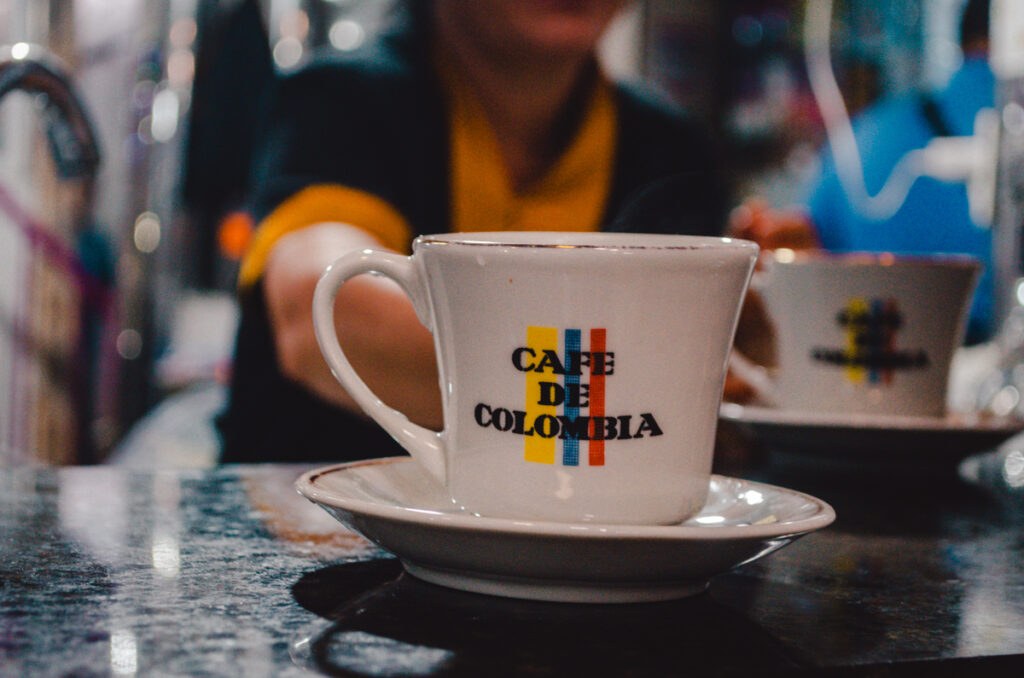 A cup of coffee with the label "Café de Colombia" on it, steaming hot, placed on a saucer on a dark table. The cup is white with a handle to the left, and the text is in the center with a simple graphic design using the colors of the Colombian flag - yellow, blue, and red. In the blurred background, a person is visible, suggesting a social setting such as a café or market. The setting appears cozy and informal, perfect for enjoying a traditional Colombian coffee.