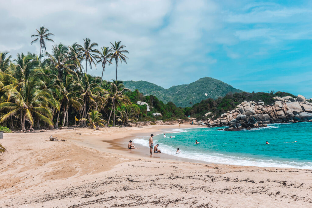 Sandy beach at Tayrona National Park with swaying coconut palm trees and visitors enjoying the turquoise sea, against a backdrop of rolling hills and scattered boulders, reflecting a tranquil tropical paradise in Colombia.