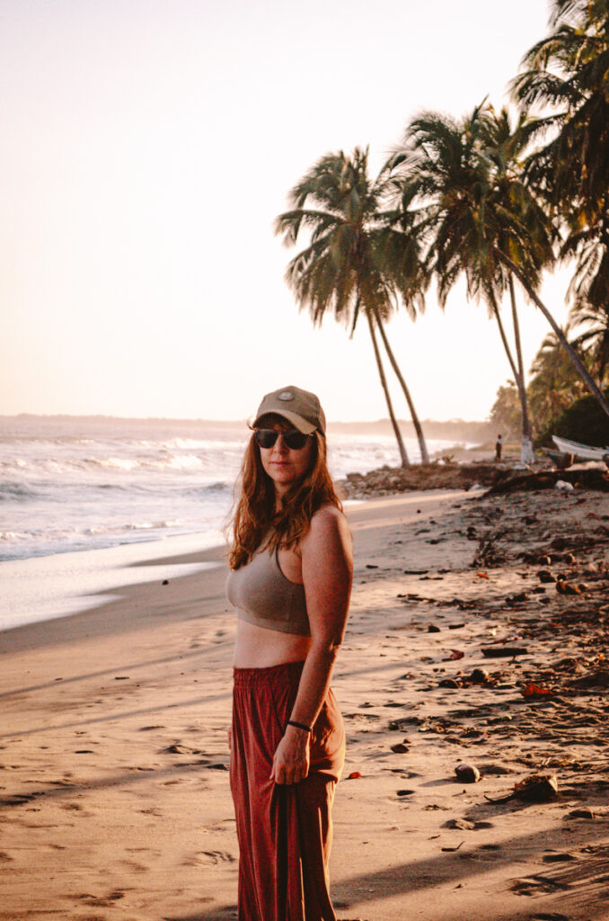 A woman stands on the sandy shores of Dibulla, Colombia, the gentle waves of the ocean in the background and a row of towering palm trees lining the beach. She is dressed for the tropical climate in a light, sleeveless top and flowing red pants, complemented by a cap. Her pose is relaxed, her face partially shaded by eco-friendly sunglasses, which reflect a commitment to protecting both her eyes and the environment around her. The setting sun casts a warm, golden light, enhancing the idyllic, peaceful ambiance.