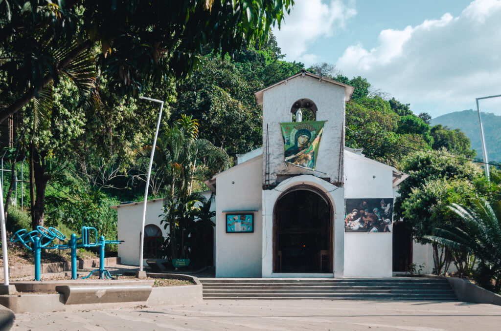 The quaint church in the center of Minca, Colombia, with a simple white facade adorned with religious artwork, surrounded by lush greenery and set against a clear blue sky.