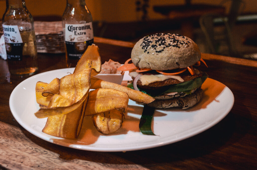 A gourmet vegan burger with a black sesame seed bun, accompanied by a side of plantain chips and a small serving of sauce, presented on a white plate with two bottles of Coronita Extra in the background.