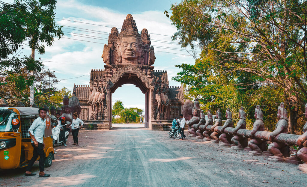 A vibrant photograph capturing the entrance to a temple with intricate designs and statues in Phnom Penh, Cambodia, known as Wat Phnom Reap. The image showcases a striking gateway adorned with traditional Cambodian sculptures and motifs, flanked by a row of guardian figures, leading to the temple grounds.