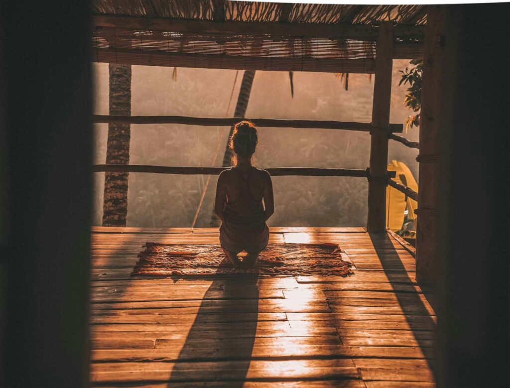 Yoga travel photo. this is a girl sitting on a mat on the wooden floor of a cabin. The sun is rising and she's doing a little yoga and meditation