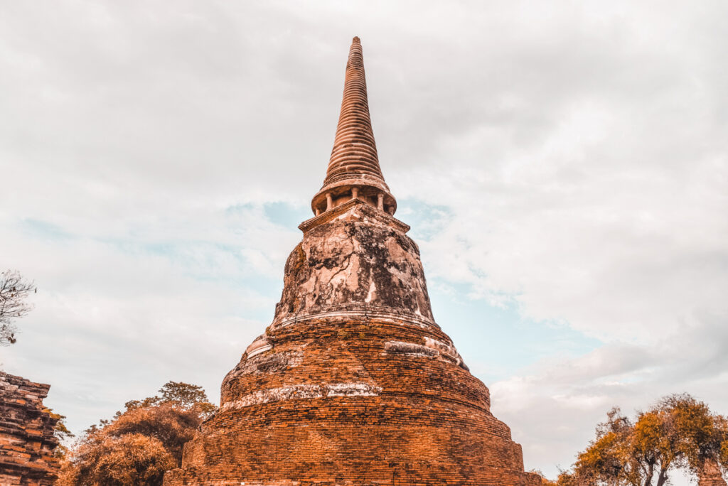 A solitary weathered brick chedi points towards the sky, surrounded by hints of greenery and under a tranquil sky, in the historic city of Ayutthaya, Thailand. This landscape encapsulates the serenity and the architectural wonder of the place, making it an ideal day trip from Bangkok to explore Thailand's rich heritage.