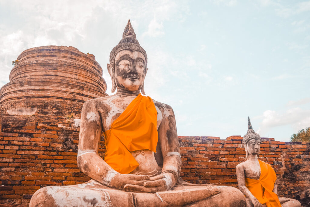 A close-up of a Buddha statue in meditation, wrapped in an orange robe against the historical backdrop of Ayutthaya's temple ruins. The statue exudes calm and spirituality, representing the city's enduring cultural significance and making it a profoundly memorable day trip from Bangkok.