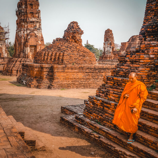 A Buddhist monk in a vibrant orange robe walks beside ancient, weathered brick ruins under a hazy sky. The ruins of Ayutthaya which can be visited on a day trip from Bangkok, stand as silent witnesses to the rich history of Thailand, with remnants of structures in the background partially enveloped in scaffolding, indicating ongoing preservation efforts. The monk's peaceful demeanor contrasts with the stark, time-worn textures of the bricks, offering a sense of continuity amidst the relics of the past.