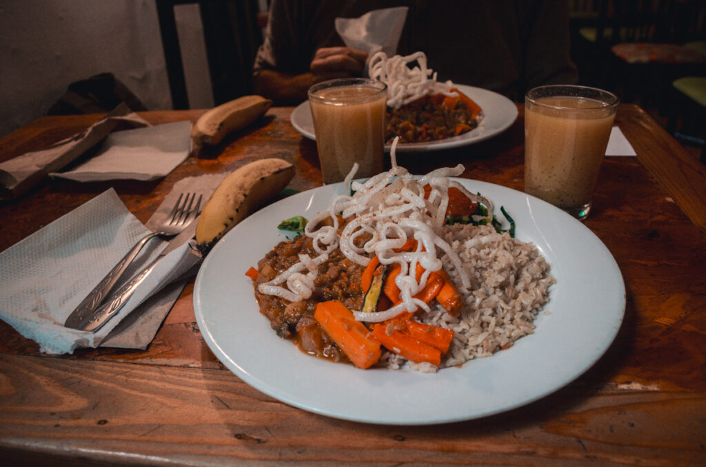 In a vegan restaurant in Bogotá, Naturalmente, there are two plates on a wooden table served with a wholesome meal. The dishes include brown rice, savory vegetable stew with carrots and other vegetables, topped with fried spaghetti, alongside a ripe banana and a smooth, tan-colored beverage. The rustic and inviting atmosphere suggests a casual dining experience focusing on plant-based cuisine.