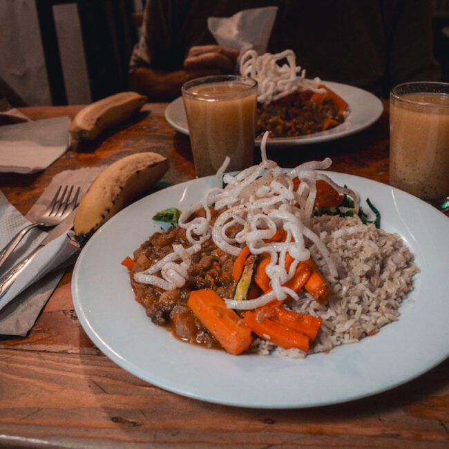 In a vegan restaurant in Bogotá, Naturalmente, there are two plates on a wooden table served with a wholesome meal. The dishes include brown rice, savory vegetable stew with carrots and other vegetables, topped with fried spaghetti, alongside a ripe banana and a smooth, tan-colored beverage. The rustic and inviting atmosphere suggests a casual dining experience focusing on plant-based cuisine.