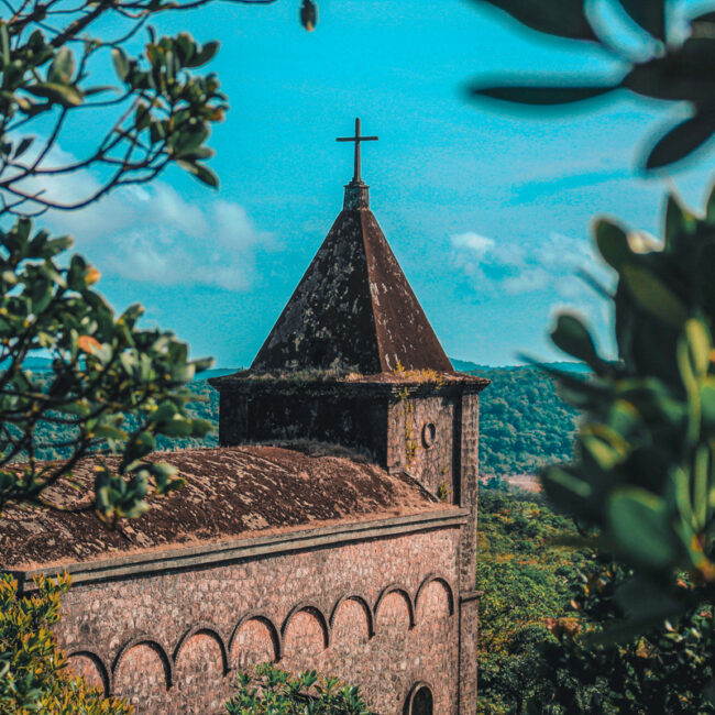 Old stone church with a pointed roof and cross atop, framed by branches with green leaves, set against a clear blue sky in Bokor Mountain Garden Center, Kampot, Cambodia.