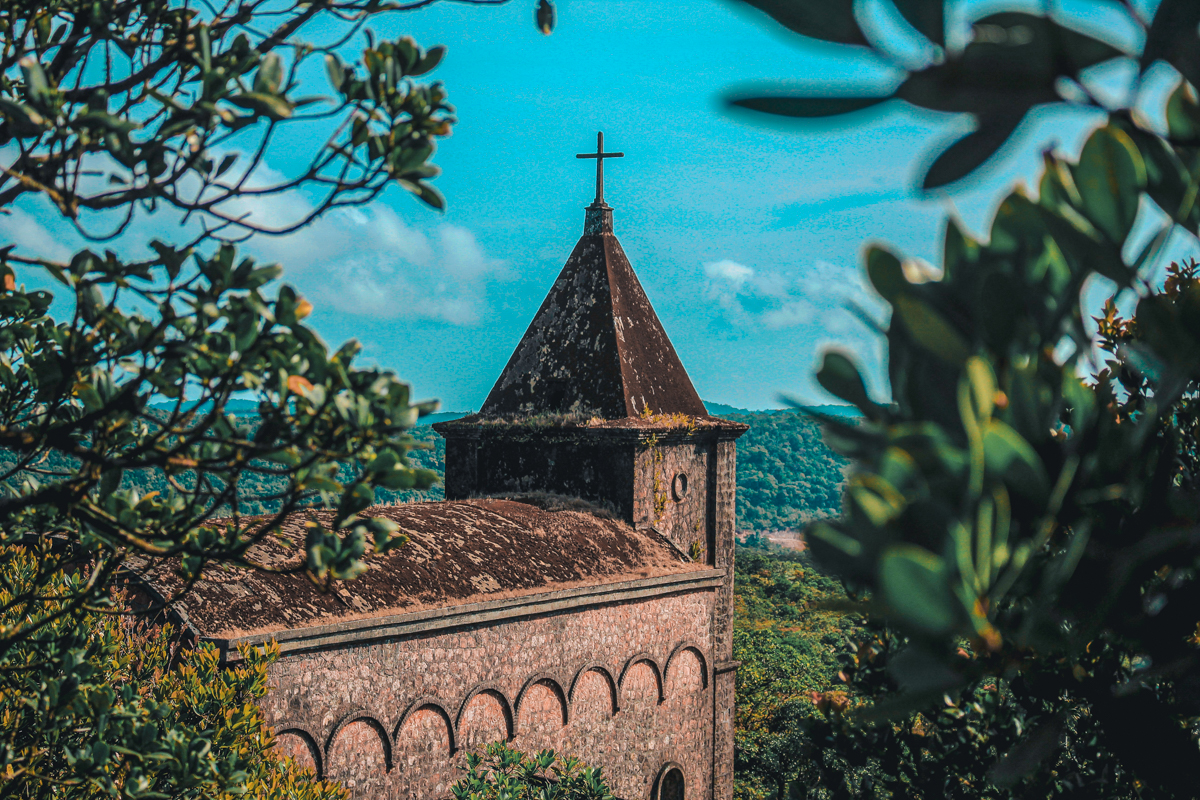 Old stone church with a pointed roof and cross atop, framed by branches with green leaves, set against a clear blue sky in Bokor Mountain Garden Center, Kampot, Cambodia.