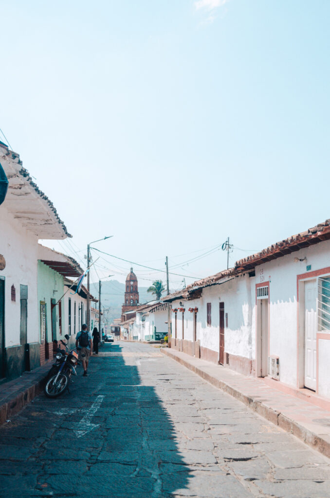 A street view in Curiti, Colombia, with a clear view of the Church of Curiti's bell tower at the end of the road, under a bright blue sky.