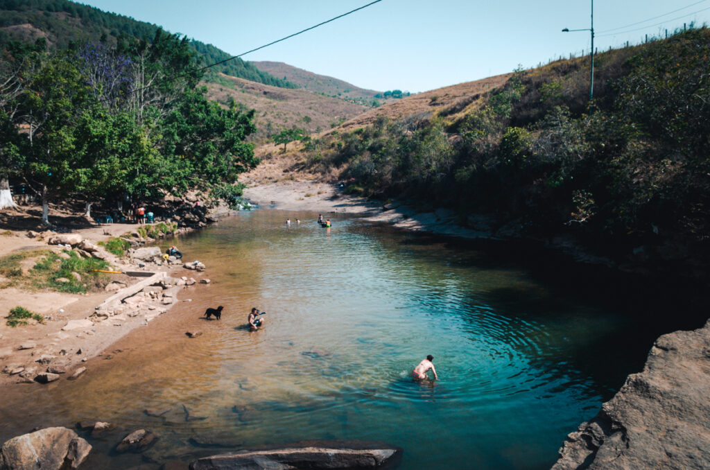 People and dogs enjoy a sunny day by the waterside at Pescaderito Curiti, Colombia, with the vibrant landscape of trees and hills in the background.