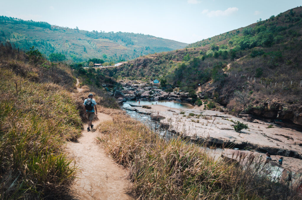 A hiker with a backpack treks along a winding trail overlooking the tranquil Pescaderito river in Curiti, Colombia, with rocky terrains and lush hills stretching into the distance.