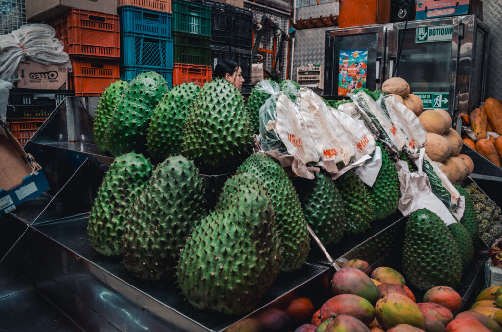 Large, spiky guanabanas (soursop) are prominently displayed at the Paloquemao market in Bogota, Colombia, with a background of mangoes and stacked crates, with a vendor in the backdrop examines the selection.