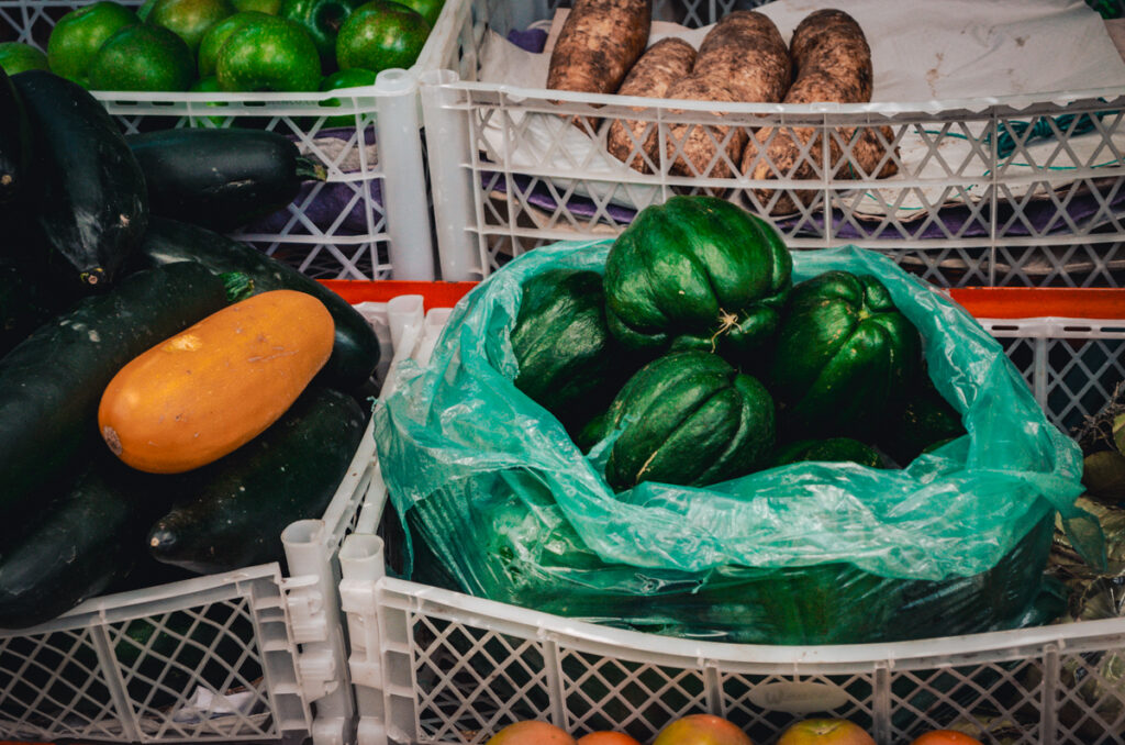 Fresh vegetables including dark green zucchinis, a lone yellow squash, and a bag of bumpy guatila, also known as chayote, displayed at Paloquemao market in Bogota, Colombia, with yams and green limes in the background.