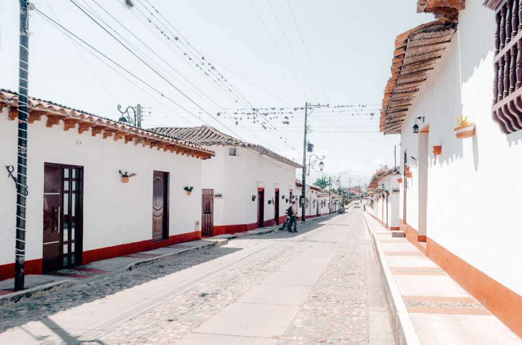 A peaceful street scene in La Playa de Belen, Colombia, featuring white colonial houses with terracotta roof tiles and decorative potted plants, under a clear blue sky.