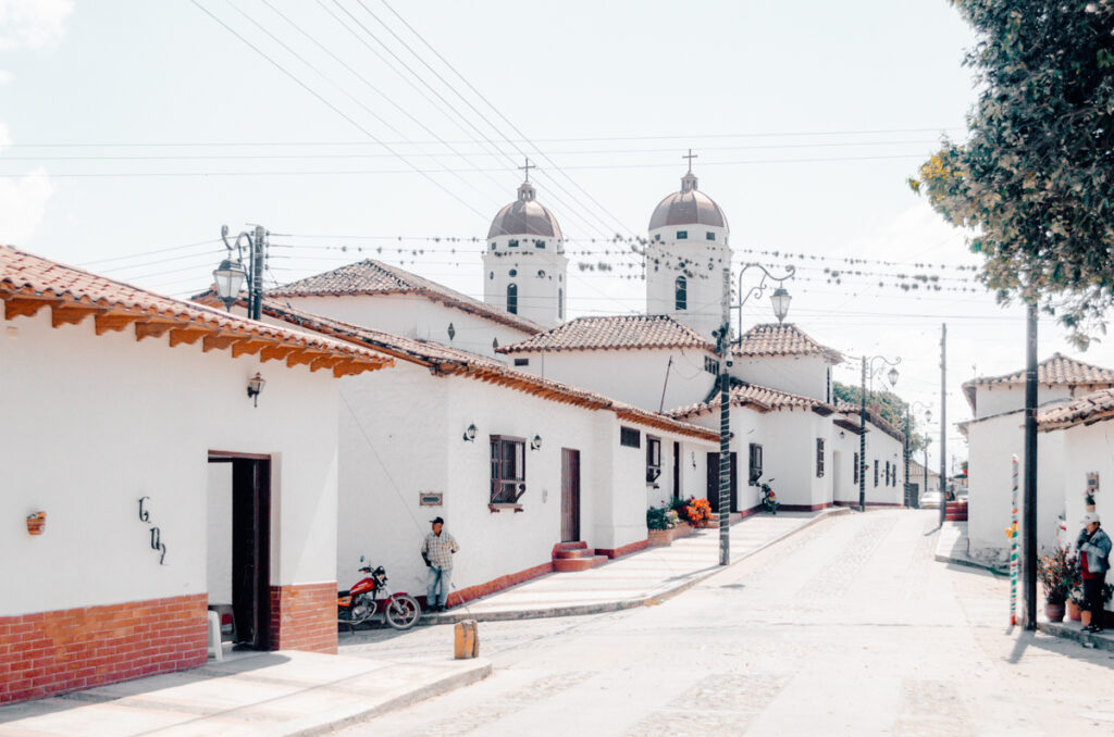 Traditional white-washed Colombian houses with terracotta tiled roofs line a cobblestone street in La Playa de Belén, with the twin domes of a church peering above the rooftops under a bright blue sky.