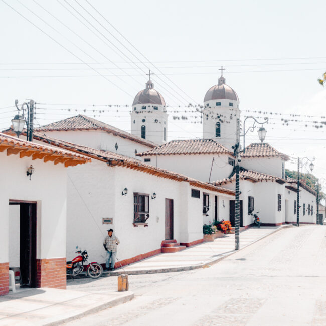 Traditional white-washed Colombian houses with terracotta tiled roofs line a cobblestone street in La Playa de Belén, with the twin domes of a church peering above the rooftops under a bright blue sky.