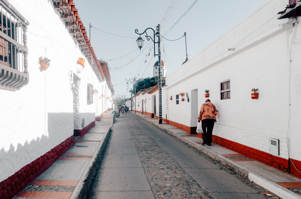 A person in an orange jacket walks down a cobblestone street lined with white colonial-style buildings in La Playa de Belen, Colombia. Potted plants add a touch of green to the sunlit scene.
