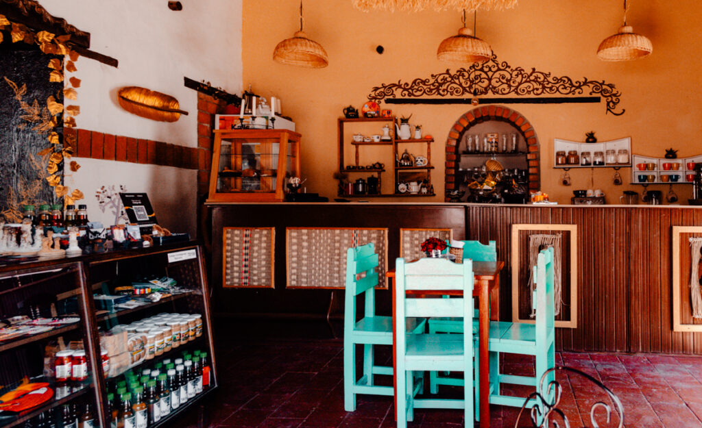 The cozy interior view of Papo Art cafe & Artesanias with vibrant turquoise chairs, rustic wooden bar, and shelves filled with various items and knick-knacks. The warm tones and artisanal decor invite a relaxing atmosphere.