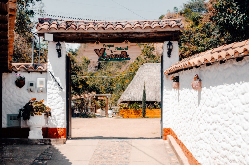 The welcoming entrance to 'Parque Natural Yaragua' with a beautifully crafted wooden sign, thatched roofing, and white walls with potted plants, inviting visitors into a space that celebrates the natural beauty of La Playa de Belen, Colombia.