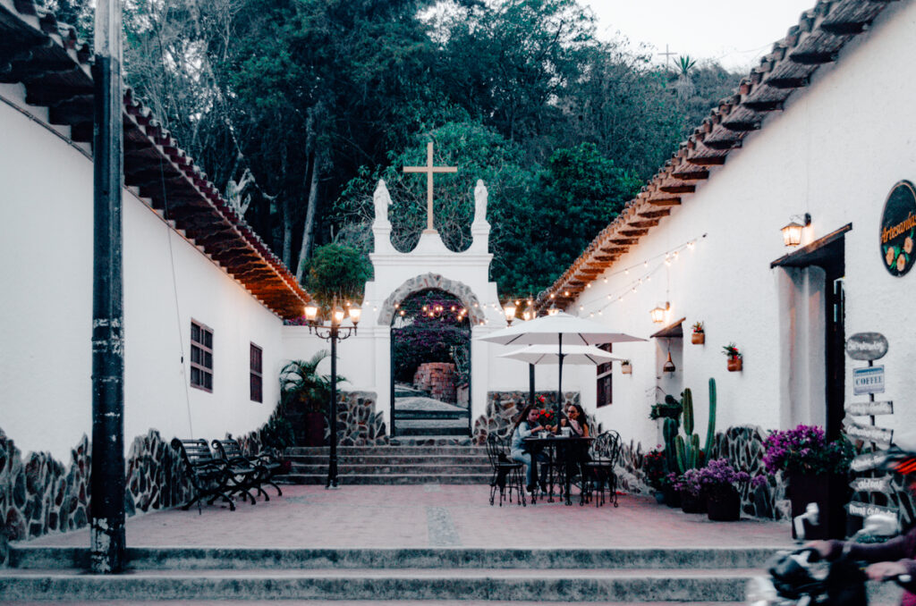 An inviting courtyard at the entrance of the cemetary at dusk with an arched entrance featuring a cross, statues, and warm glowing lights. Two people sit at a table under an umbrella, creating a serene and intimate setting in La Playa de Belen, Colombia.