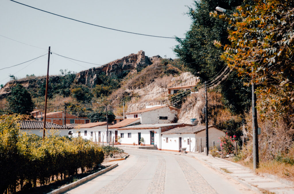 A cobblestone street in La Playa de Belen, Colombia, meanders gently past white-walled homes with terracotta roofs, leading towards a striking hillside that looms in the background, illustrating the town’s harmony with its natural surroundings.