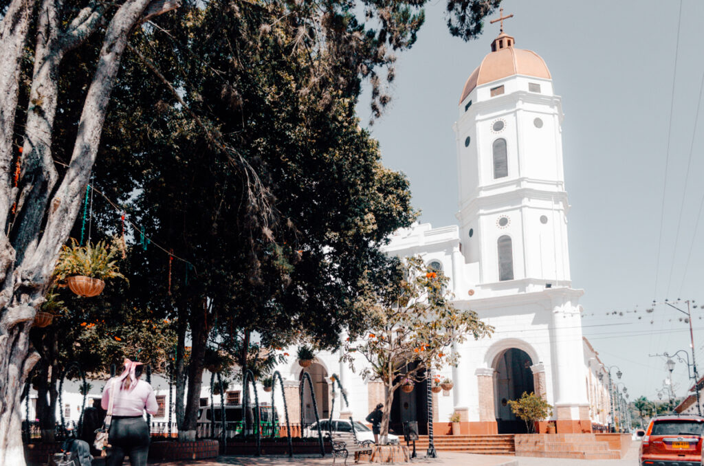 A sunny view of the whitewashed bell tower of a church in La Playa de Belen, Colombia, with a foreground of lush trees and a person walking towards the entrance. A festive atmosphere is hinted at by colorful hanging decorations amidst the greenery.