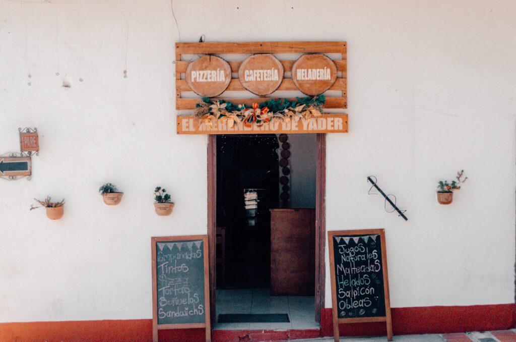 The rustic facade of 'El Merendero de Yader,' a local establishment in La Playa de Belen offering pizzeria, cafeteria, and ice cream services. The entrance is adorned with wooden signage and flanked by chalkboards listing beverages and snacks such as 'jugos naturales' and 'helados'.