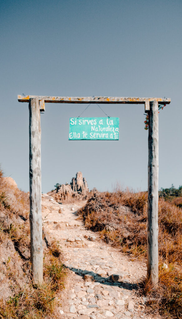 A rustic wooden sign with the message 'Si sirves a la Naturaleza ella te servirá a ti' (If you serve nature, it will serve you) stands at the entrance of a trail leading towards towering clay formations in Los Estoraques Unique Natural Area, against a clear blue sky.