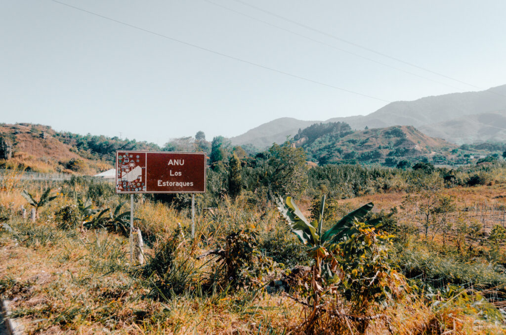 Signage reading 'ANU Los Estoraques' alongside a rural road with a background of lush greenery and rolling hills leading to Los Estoraques Unique Natural Area, under a bright blue sky.