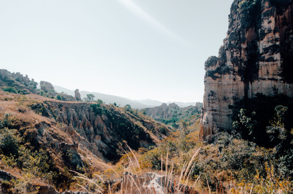 A serene landscape view of Los Estoraques Unique Natural Area, featuring a deep valley with eroded clay formations surrounded by lush vegetation and a tall cliff face under a clear sky with a ray of sunlight.