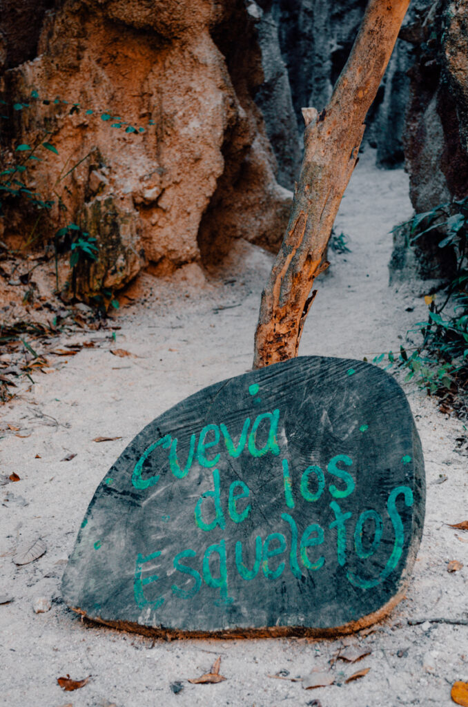 A hand-painted sign on a stone slab reads 'Cueva de los Esqueletos' (Cave of Skeletons) at the entrance of a narrow pathway flanked by high eroded walls in Los Estoraques Unique Natural Area.